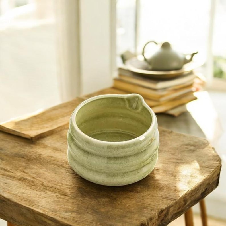 May include: A light green ceramic matcha bowl with a textured, swirling design. The bowl has a slight lip on one side and is sitting on a wooden surface.