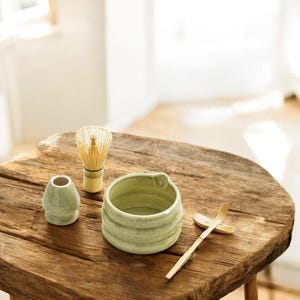 May include: A green ceramic matcha bowl, a bamboo whisk, a green ceramic teacup, and a bamboo matcha scoop on a wooden table.