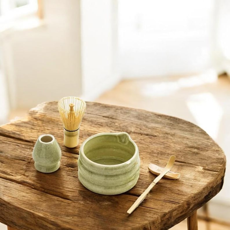 May include: A green ceramic matcha bowl, a bamboo whisk, a green ceramic teacup, and a bamboo matcha scoop on a wooden table.