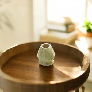May include: A light green ceramic matcha whisk holder on a wooden tray. The holder has a rounded base and a wide opening at the top.