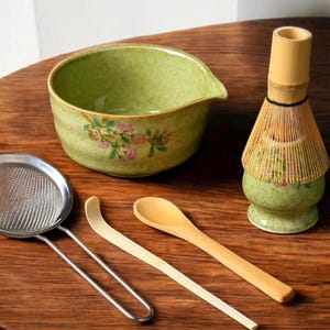 May include: A green ceramic matcha bowl with floral designs, a bamboo whisk with a green ceramic base, a stainless steel strainer, a wooden spoon, and a white bamboo matcha scoop.