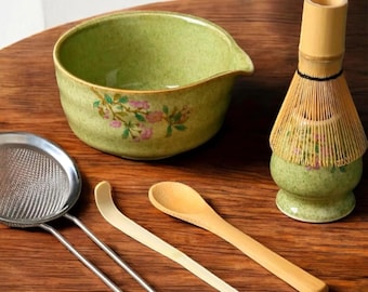 May include: A green ceramic matcha bowl with floral designs, a bamboo whisk with a green ceramic base, a stainless steel strainer, a wooden spoon, and a white bamboo matcha scoop.