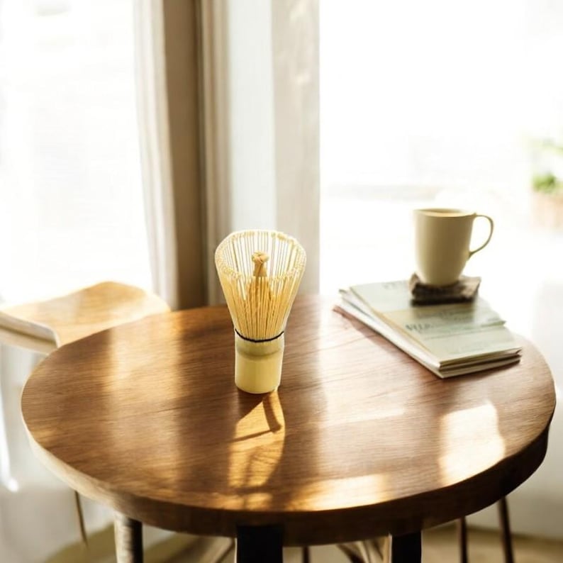 May include: A wooden table with a natural finish, a white ceramic mug, and a bamboo matcha whisk. The whisk is standing upright on the table.