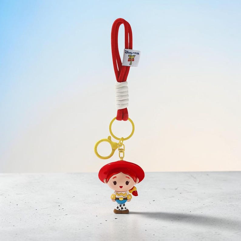 Puede incluir: Un cord&oacute;n rojo con una secci&oacute;n envuelta en blanco y una peque&ntilde;a etiqueta blanca con el logotipo de Disney Pixar Toy Story 3. Unido al cord&oacute;n hay un mosquet&oacute;n y una anilla de pl&aacute;stico amarillo que sostienen una figura de una vaquera.