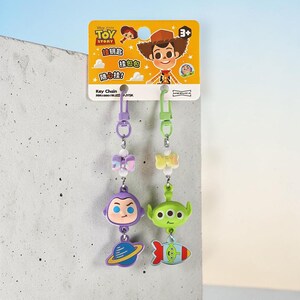 Puede incluir: Dos llaveros de Toy Story en una tarjeta amarilla. Uno presenta una cabeza morada de Buzz Lightyear y un dije de planeta. El otro presenta una cabeza verde de Alien y un dije de cohete. Ambos tienen un peque&ntilde;o lazo iridiscente y un mosquet&oacute;n.