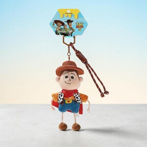 Puede incluir: Un llavero de peluche de Woody de Toy Story 5 con sombrero de vaquero marr&oacute;n, chaleco amarillo con estampado de vaca, pa&ntilde;uelo rojo y pantalones azules. El personaje est&aacute; unido a una correa de mu&ntilde;eca trenzada marr&oacute;n y una tarjeta de cart&oacute;n.