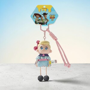 Puede incluir: Un llavero de peluche de Bo Peep con un personaje rubio en un vestido azul claro con un lazo rosa, unido a un cord&oacute;n rosa. El encabezado del empaque muestra el logo de Toy Story 5 e ilustraciones de Jessie, Buzz Lightyear y Woody.