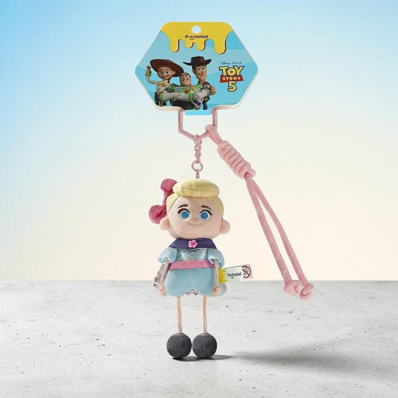Puede incluir: Un llavero de peluche de Bo Peep con un personaje rubio en un vestido azul claro con un lazo rosa, unido a un cord&oacute;n rosa. El encabezado del empaque muestra el logo de Toy Story 5 e ilustraciones de Jessie, Buzz Lightyear y Woody.