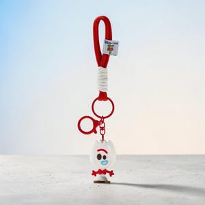 Puede incluir: Un llavero de cord&oacute;n rojo y blanco con un peque&ntilde;o dije de pl&aacute;stico blanco que representa un personaje con una boca roja, un ojo negro y un ojo azul. Incluye una etiqueta blanca con el texto Disney Pixar Toy Story 5 y una argolla roja.