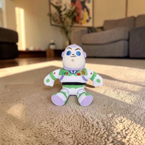 May include: A plush toy character featuring a white, green, and purple space ranger suit with a gold star badge on the chest. The figure has a rounded head, blue eyebrows, and a small chin detail, designed with a soft, stuffed fabric construction.