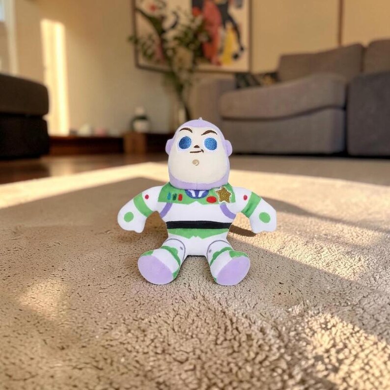May include: A plush toy character featuring a white, green, and purple space ranger suit with a gold star badge on the chest. The figure has a rounded head, blue eyebrows, and a small chin detail, designed with a soft, stuffed fabric construction.