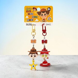 Puede incluir: Dos llaveros de Toy Story en una tarjeta amarilla. Uno presenta la cabeza del personaje Woody con una estrella de sheriff amarilla, y el otro la cabeza de Jessie con un sombrero rojo. Ambos tienen clips de metal dorado y rojo.