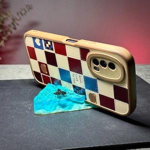 May include: A tan phone case with a red, blue, and white checkered pattern, featuring small bear illustrations and the text "Good Luck To You." The case is propped up by a blue resin stand resembling water.
