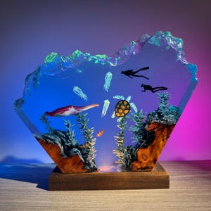 May include: A handcrafted resin sculpture depicting an underwater scene. The artwork features a squid, sea turtle, jellyfish, and divers. The base is wood, and the resin is blue and clear, with a colorful gradient background.