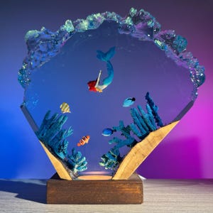 May include: A decorative resin sculpture featuring a mermaid with red hair and a blue tail swimming in a clear, blue-toned environment. The sculpture includes coral, fish, and a wooden base. The top edge has a textured, crystal-like appearance.