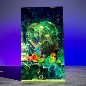 May include: A rectangular resin art piece featuring an underwater scene. The artwork showcases colorful coral, jellyfish, and a rocky background. The piece is illuminated with green and blue hues, creating a vibrant, aquatic display.