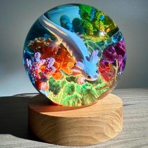 May include: A decorative resin sphere featuring a light blue axolotl swimming among colorful coral and plants. The sphere is mounted on a round, light-colored wooden base, creating a unique art piece.