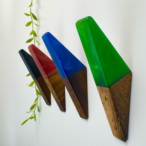 May include: Four wall hooks with a geometric design. Each hook features a wooden base and a colorful resin top in black, red, blue, and green. The hooks are mounted on a white wall, with a green leafy vine in the background.