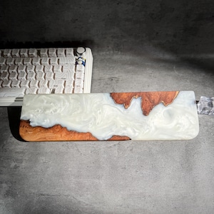 May include: A rectangular wooden wrist rest with a white and brown resin design. The wrist rest is designed to be used with a keyboard. The wood has a natural grain pattern, and the resin has a marbled effect.