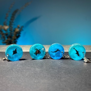 May include: Four round, blue resin keychains with sea creature designs. Each keychain features a different black silhouette: a manta ray, a sea turtle, a diver, and a shark. The keychains have silver-colored hardware.