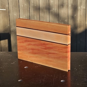 Puede incluir: Una tabla de cortar rectangular de madera con un diseño multicolor. La sección superior es de color marrón claro, seguida de una fina franja marrón oscuro, una sección beige, otra franja marrón oscuro y una base marrón rojizo. La tabla está de pie.