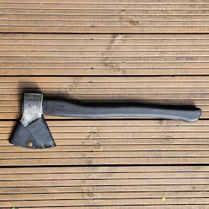 May include: A hand axe featuring a dark, textured wooden handle and a metallic head with a weathered finish. The blade is partially covered by a black leather sheath secured with a single brass-colored metal snap button and two rivets.