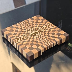 Puede incluir: Tabla de cortar cuadrada de madera con un diseño de ilusión óptica único. La tabla presenta un patrón de tablero de ajedrez que converge hacia el centro, creando un efecto 3D. Los tonos de madera incluyen cuadrados marrones claros y oscuros.
