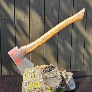 May include: A metal axe with a weathered, red-painted blade and a curved, light brown wooden handle. The axe is embedded into a rough, cylindrical piece of tree trunk featuring textured bark and patches of yellow lichen on its surface.