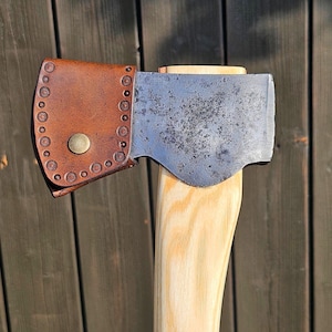 May include: Close-up of a vintage axe with a wooden handle and a metal blade. The axe head is covered by a brown leather sheath with decorative stitching and a brass button closure. The wooden handle is light-colored.