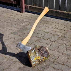 Könnte beinhalten: Ein Axtkopf aus Stahl mit einem geschwungenen Holzstiel, der in einem zylindrischen Holzstamm steckt. Die Axt hat eine metallisch graue Klinge und einen hellen, glatten Holzstiel. Der Stamm hat eine raue Rinde mit gelben Flechten.