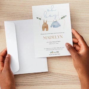 Puede incluir: Una invitación blanca para baby shower con el texto "oh boy!" en escritura azul. La invitación presenta ilustraciones de acuarela de ropa de bebé colgada en un tendedero. La invitación incluye el nombre "Madelyn" y los detalles del evento.