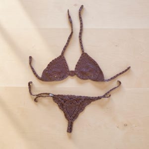 Peut inclure: Ensemble de bikini en crochet marron. Le haut est de style triangle avec de longues attaches, et le bas est de style string. L'ensemble est fait d'un fil doux et texturé, présenté sur une surface en bois clair.