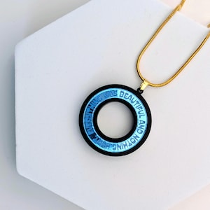 Könnte beinhalten: Eine schwarze und blaue kreisförmige Anhänger-Halskette mit den Worten "EVERYTHING WAS BEAUTIFUL AND NOTHING HURT" in weißer Schrift. Der Anhänger hängt an einer goldfarbenen Kette.