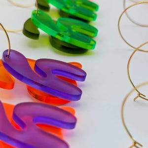 Peut inclure: Gros plan sur des boucles d'oreilles en acrylique colorées. Les boucles d'oreilles présentent un design abstrait unique dans les tons de violet, d'orange et de vert. Des créoles dorées sont également visibles. Les boucles d'oreilles sont présentées sur une surface blanche.