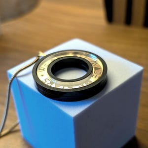 May include: A black and gold pendant necklace with a circular design. The pendant has a clear center with gold lettering and a black outer ring. The necklace is displayed on a light blue cube.