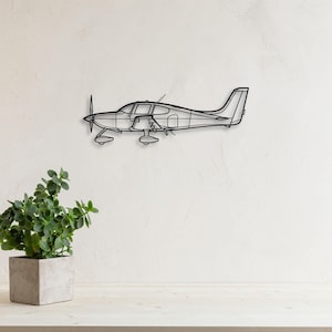 May include: Black metal airplane wall art. The airplane features a propeller, wings, and a detailed fuselage. The artwork is mounted on a white wall, with a potted plant in the foreground.