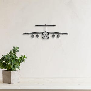 May include: A black metal airplane wall art piece. The airplane has a detailed design with wings, a tail, and multiple wheels. The artwork is mounted on a white wall, with a potted plant in the foreground.