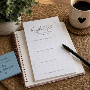 May include: A white notepad titled Messy Mind Pad with prompts for thoughts, resting on a spiral notebook. A blue sticky note with handwritten tasks, a black pen, and a white mug with a black heart are present. The notepad features floral line art.