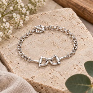 May include: A silver-toned chain link bracelet featuring a central equestrian-inspired bit motif and a circular toggle clasp closure. The bracelet is constructed from polished metal links that form a continuous circular band around the wrist.