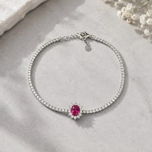 May include: A silver-toned tennis bracelet featuring a continuous row of small clear stones. The center of the bracelet showcases a prominent oval-shaped vibrant pink stone surrounded by a halo of smaller clear stones and a small metal tag.