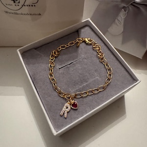 Può includere: Bracciale a catena dorata con un ciondolo a forma di lettera "R" incrostata di cristalli trasparenti e un piccolo ciondolo a forma di cuore rosso. Il bracciale è presentato in una scatola bianca con interno grigio.