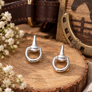May include: A pair of polished silver-tone stud earrings shaped like equestrian horse bits. The earrings feature a smooth, metallic finish with a classic snaffle bit design, consisting of a central bar connected to two circular rings on each side.