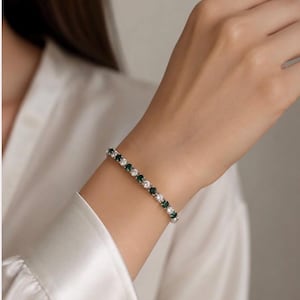 Può includere: Un bracciale tennis caratterizzato da un motivo ripetuto di pietre rotonde bianche trasparenti alternate a pietre rotonde verde smeraldo intenso, incastonate in una fascia di metallo color argento lucido che avvolge il polso con una chiusura.