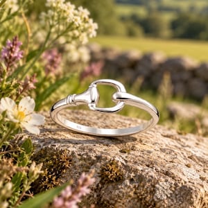 May include: A polished silver ring featuring a horse bit design. The band is smooth and rounded, with a central horizontal bar and two side loops that mimic the shape of an equestrian bit. The interior of the band is clearly stamped with 925.