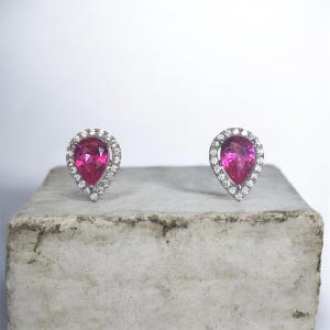 Può includere: Un paio di orecchini a lobo con una gemma centrale a forma di pera di colore rosa vivace, circondata da un alone di piccole pietre chiare e scintillanti incastonate in una montatura in metallo color argento dal design elegante.