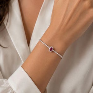 Könnte beinhalten: Ein zartes silberfarbenes Armband mit einem einzelnen ovalen, tiefroten Edelstein in der Mitte, der von einem Kranz aus kleinen klaren Steinen umgeben ist. Das Band besteht aus einer durchgehenden Reihe kleiner, funkelnder Steine in einer dünnen Kette.