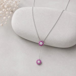 May include: A silver lariat necklace featuring two flower-shaped pendants. Each pendant is composed of a central clear stone surrounded by a ring of small, vibrant pink stones. The delicate chain connects the two floral elements in a vertical drop.