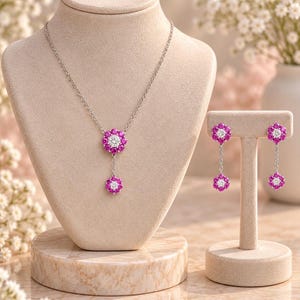 Peut inclure: Un ensemble collier et boucles d'oreilles pendantes en argent avec des motifs floraux. Chaque pièce est ornée d'une pierre centrale transparente entourée d'un halo de pierres roses vives. Le collier présente un pendentif à deux fleurs.