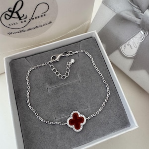 May include: A silver bracelet with a four-leaf clover charm. The clover is red with a white border and is set in a silver chain. The bracelet is presented in a white gift box with a gray interior.