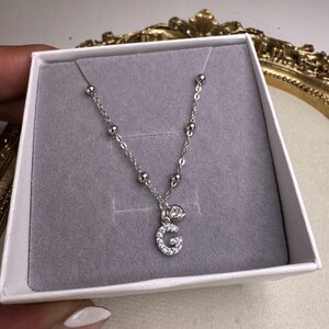 Può includere: Collana d'argento con ciondolo in una scatola bianca. La collana presenta una catena con piccole perle e un ciondolo a forma di lettera "G" incrostato di piccole pietre trasparenti. È presente anche un piccolo ciondolo a forma di cuore.
